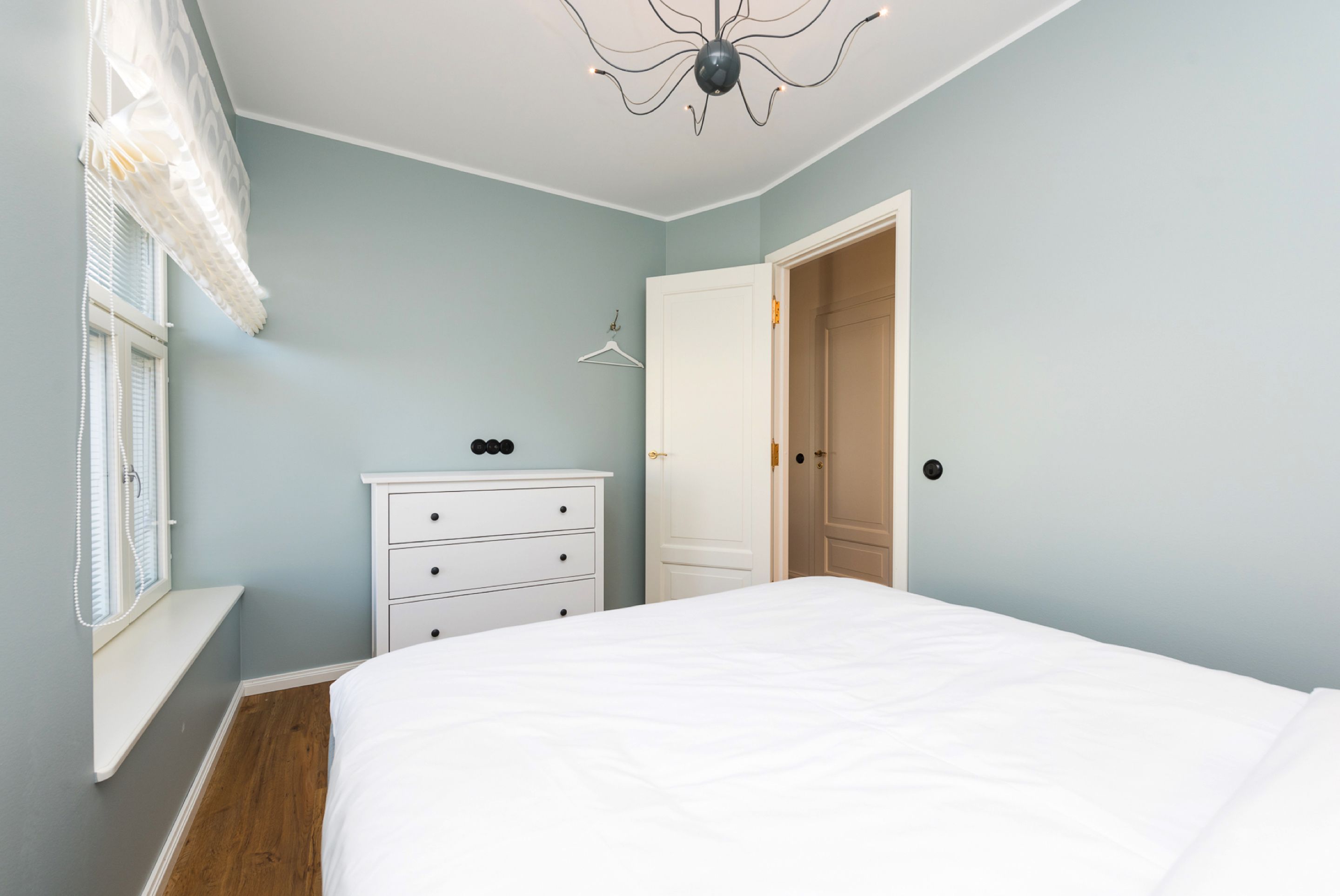 Een slaapkamer met een bed, een dressoir, een raam met zonwering en een deur.