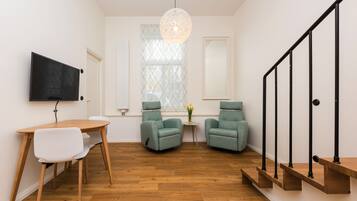 Una sala de estar moderna con suelo de madera, una mesa redonda, dos sillones verdes y un televisor negro montado en la pared.