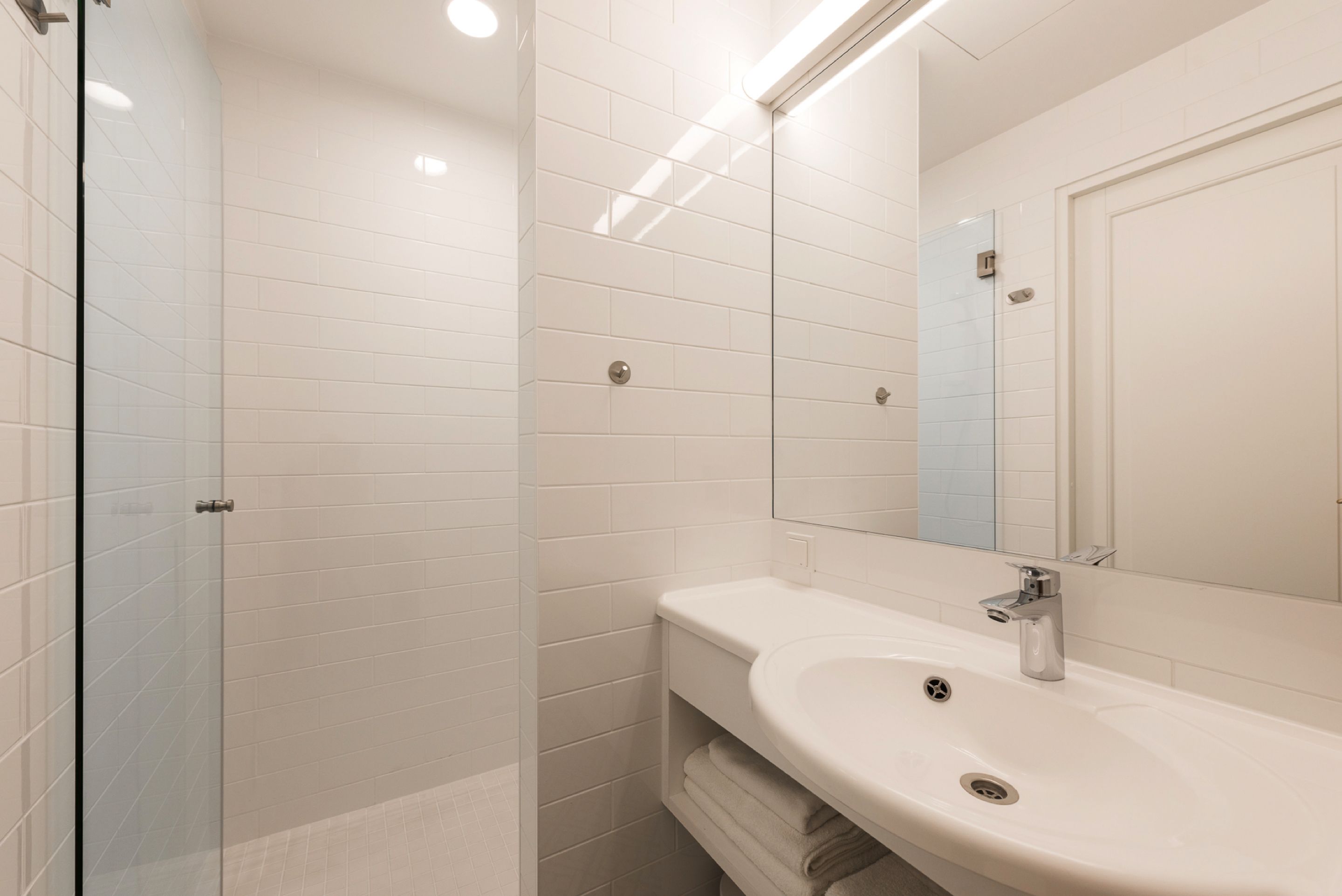 A modern bathroom with white tiles, a glass shower enclosure, a sink with a mirror, and a towel rack.