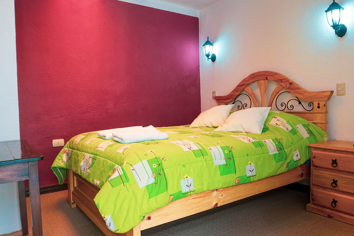 Ein Schlafzimmer mit einem hölzernen Bett, floralem Bettzeug in Grün, einem Nachttisch und einer roten Akzentwand.
