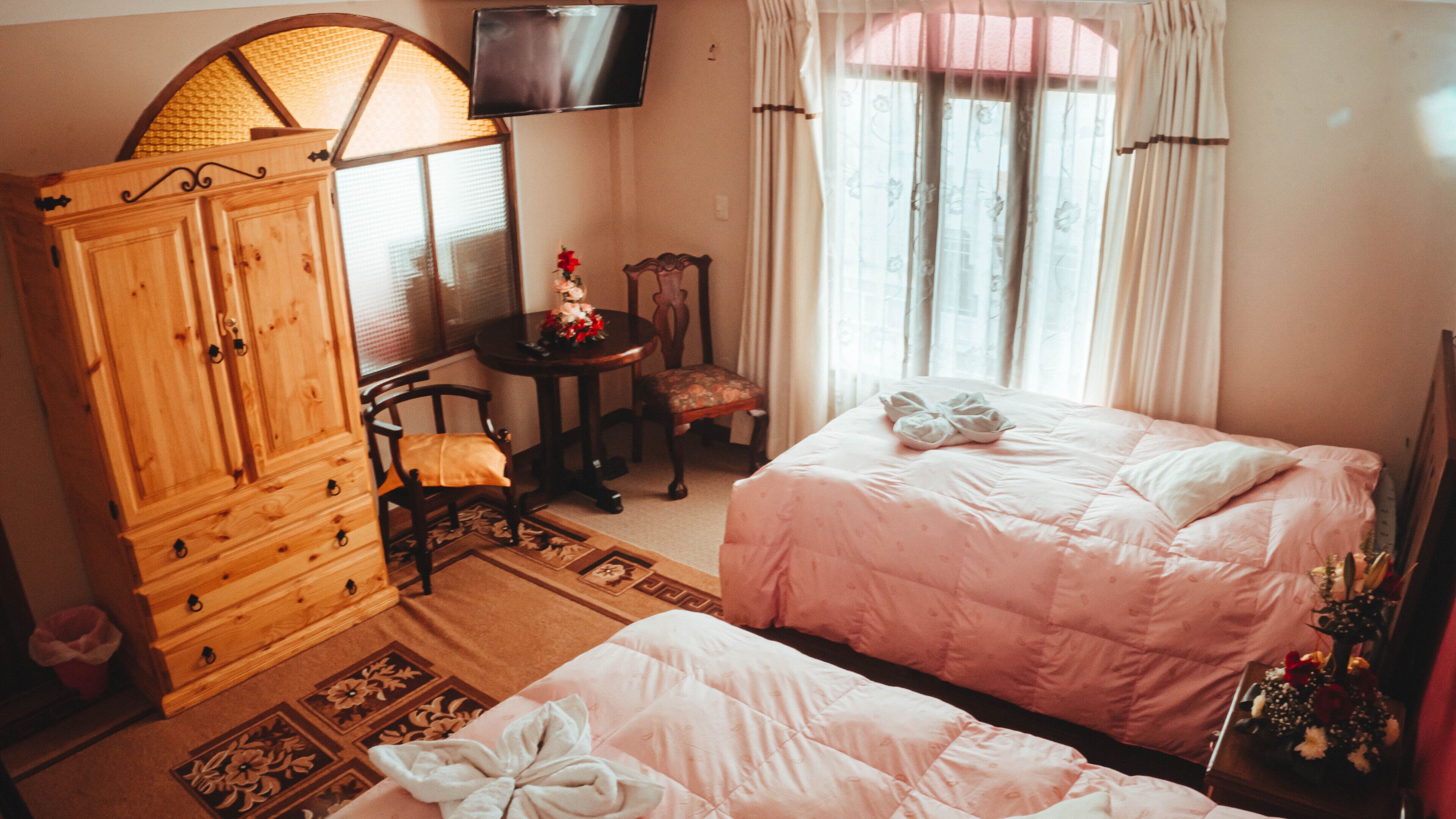 Un dormitorio con una cama, un armario de madera, una mesita con una silla, un televisor montado en la pared y una ventana con cortinas.