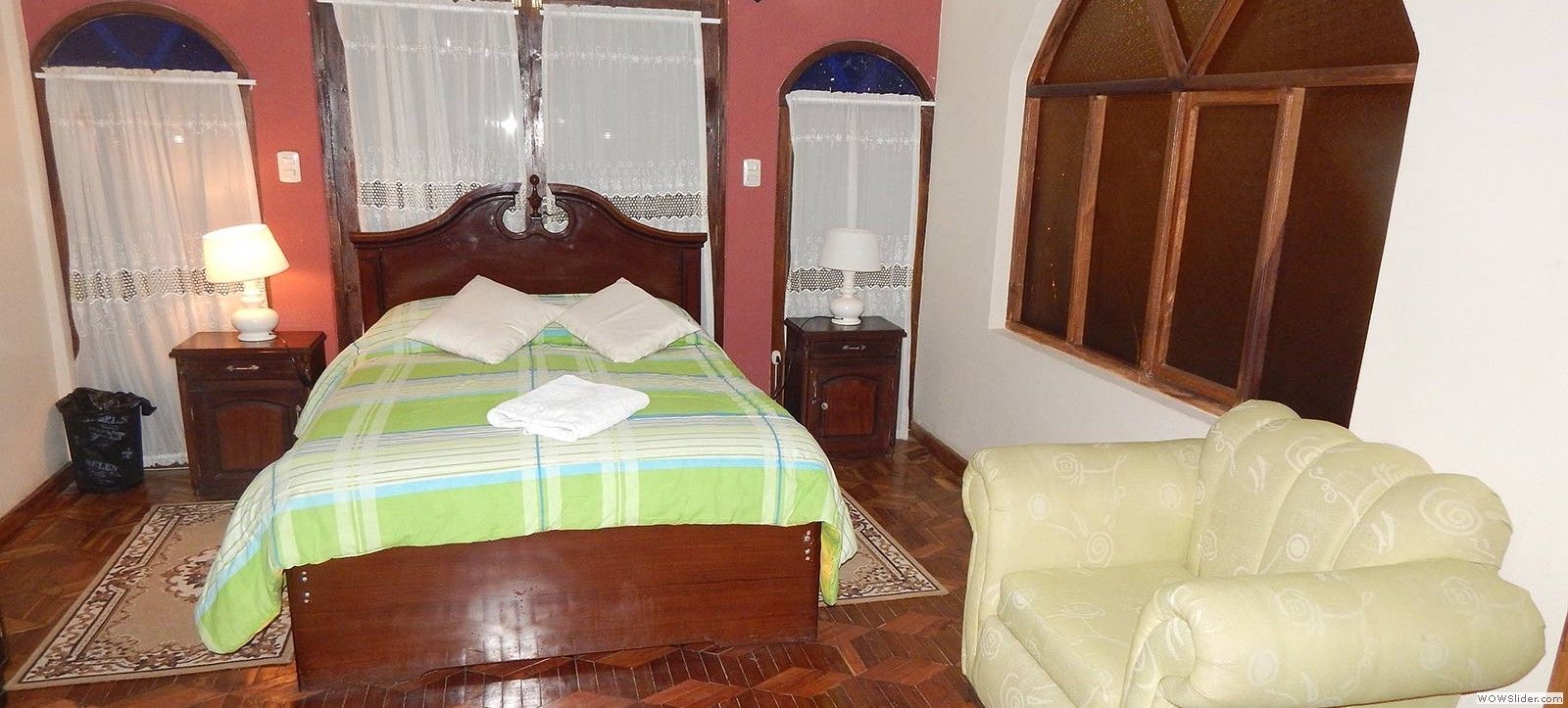 Un dormitorio con una cama de madera, dos mesitas de noche con lámparas, una ventana con cortinas y un sillón estampado.