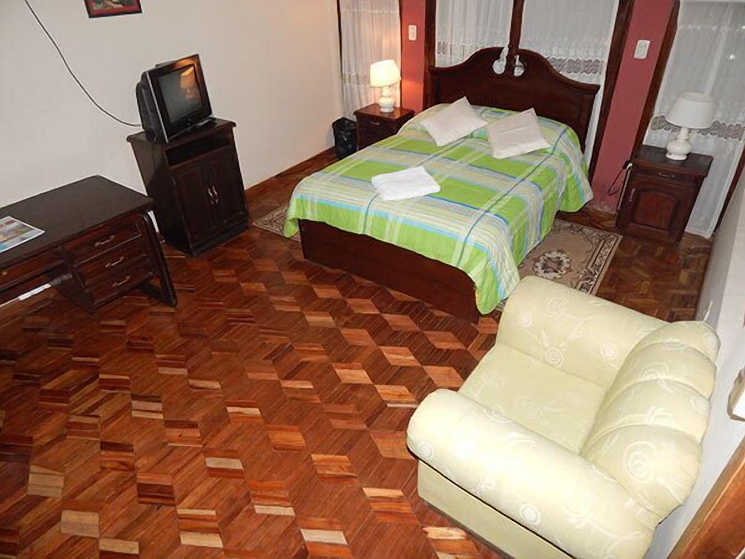 A bedroom with a wooden herringbone floor, a bed with a green and white striped bedspread, a wooden nightstand with a lamp, a television on a wooden cabinet, and a cream-colored armchair.