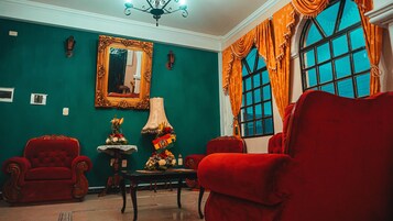 Una habitación con paredes verde agua, un espejo grande, dos sillones rojos, una mesa con una lámpara y una ventana con cortinas anaranjadas.