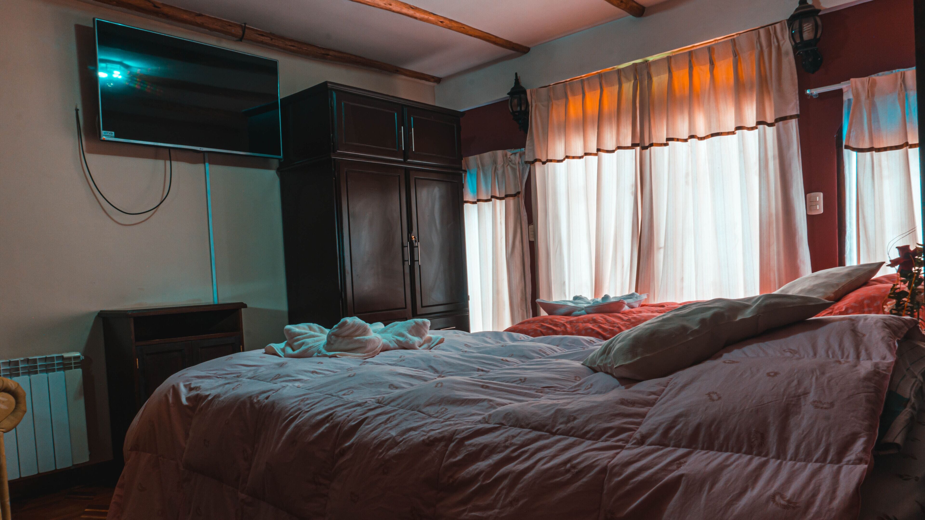 Une chambre à coucher comprenant un lit, une armoire, un téléviseur fixé au mur et une fenêtre avec des rideaux.