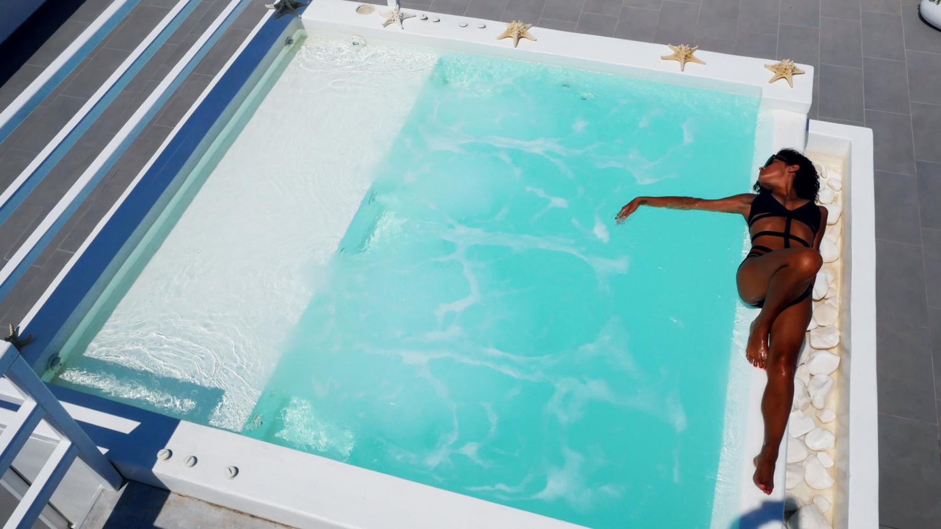 Uma pessoa deitada em uma piscina pequena e retangular, com água cristalina, cercada por uma borda de piscina branca e um deck revestido de azulejos.