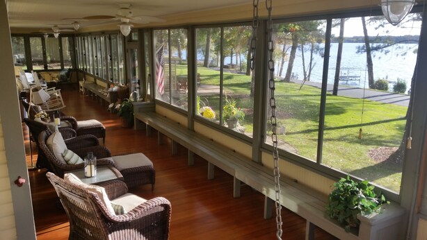 A sunlit room with wicker furniture, large windows, and a view of a lake. - Lake Ripley Lodge w Lake Front Rooms, Grand Porch, Kayaks & Paddleboard (Cambridge)