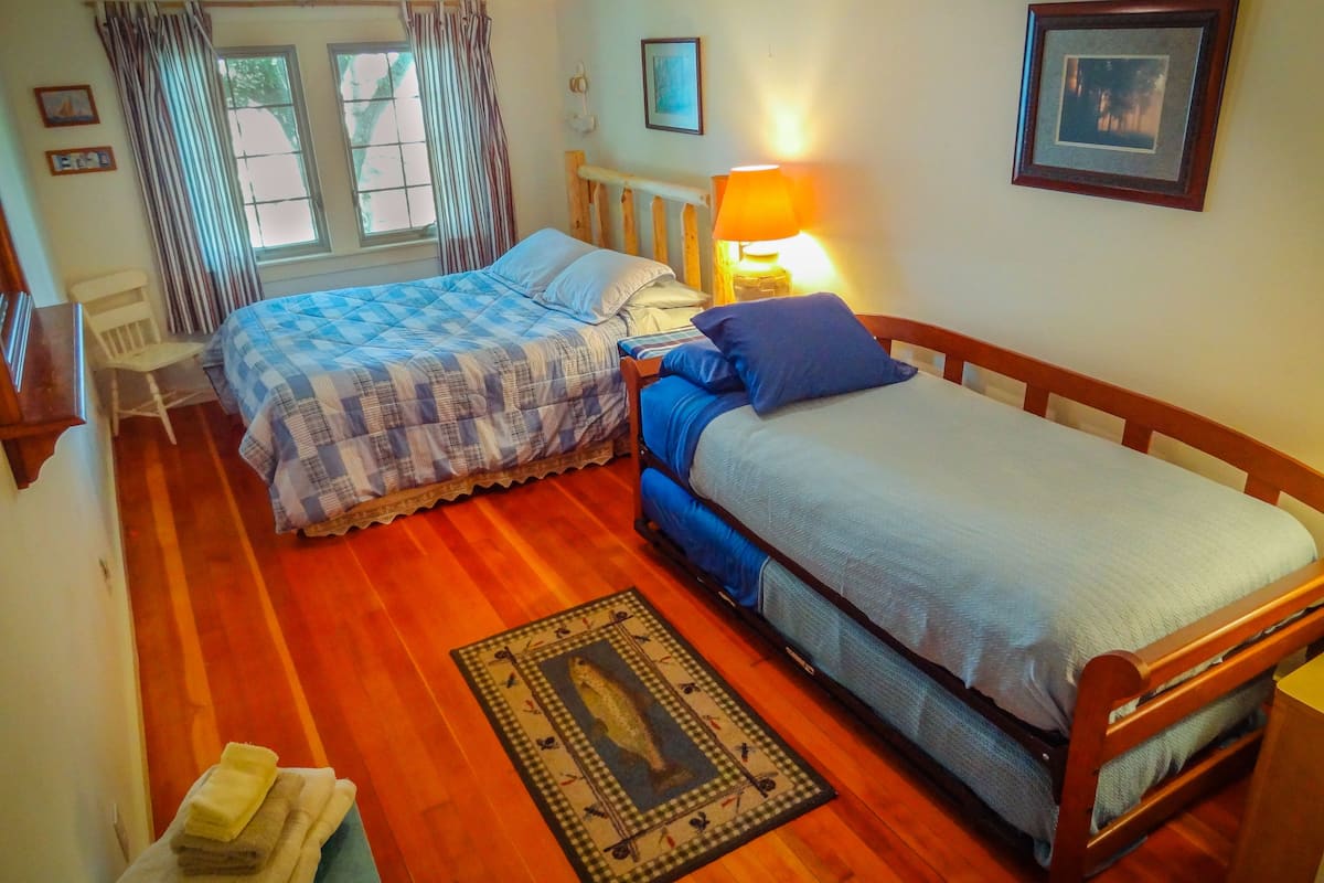 A bedroom with two beds, a wooden floor, a window with curtains, a bedside lamp, and a framed picture on the wall.