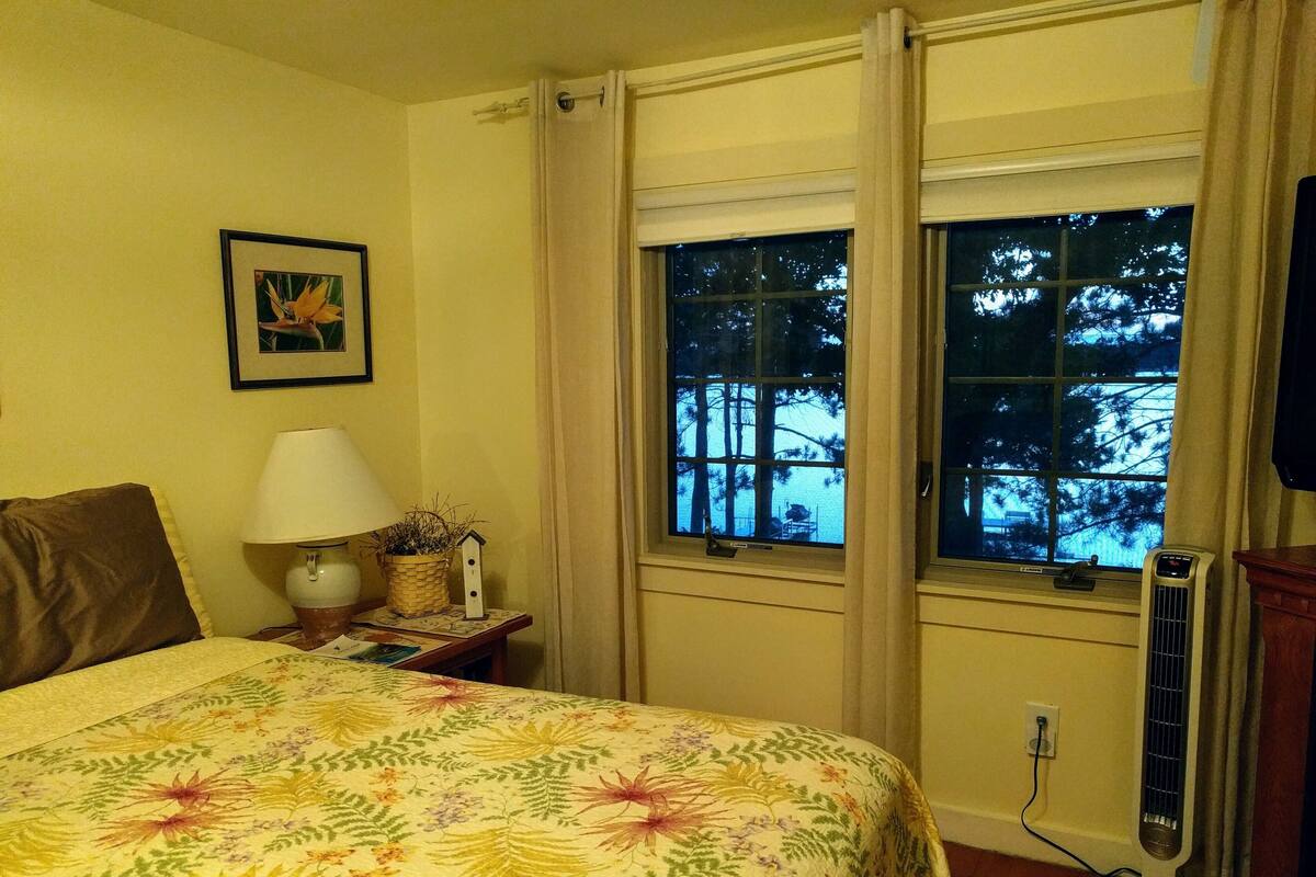 A hotel room with a bed, a nightstand with a lamp, a window with a view of trees, and a wall-mounted air conditioner.