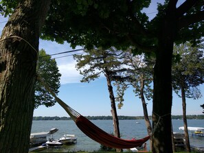 A hammock tied between trees overlooking a lake with boats docked nearby. - Lake Ripley Lodge w Lake Front Rooms, Grand Porch, Kayaks & Paddleboard (Cambridge)