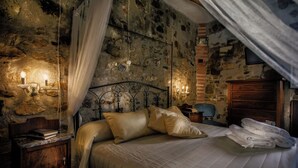 Quarto com parede de pedra, cama com roupa de cama branca, mesa de cabeceira com livros e cômoda de madeira.