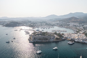 Vista aérea de una ciudad costera con una imponente estructura similar a un castillo, rodeada de agua, barcos y un puerto deportivo.