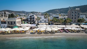 A coastal hotel with outdoor seating and umbrellas, a beach in the foreground, and buildings in the background. - Artemis Hotel (Bodrum)