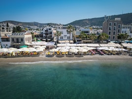 Un hotel frente al mar con terraza y sombrillas, una playa en primer plano y edificios al fondo.