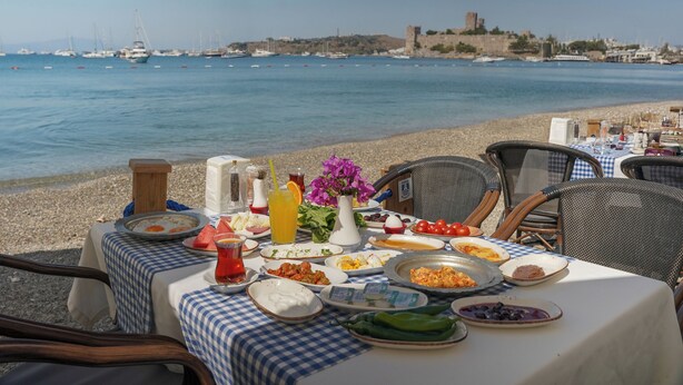 A table set on a beach with food and drinks, overlooking a calm sea and a historic building in the background. - Artemis Hotel (Bodrum)