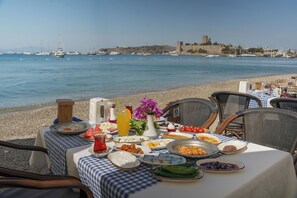 A table set on a beach with food and drinks, overlooking a calm sea and a historic building in the background.