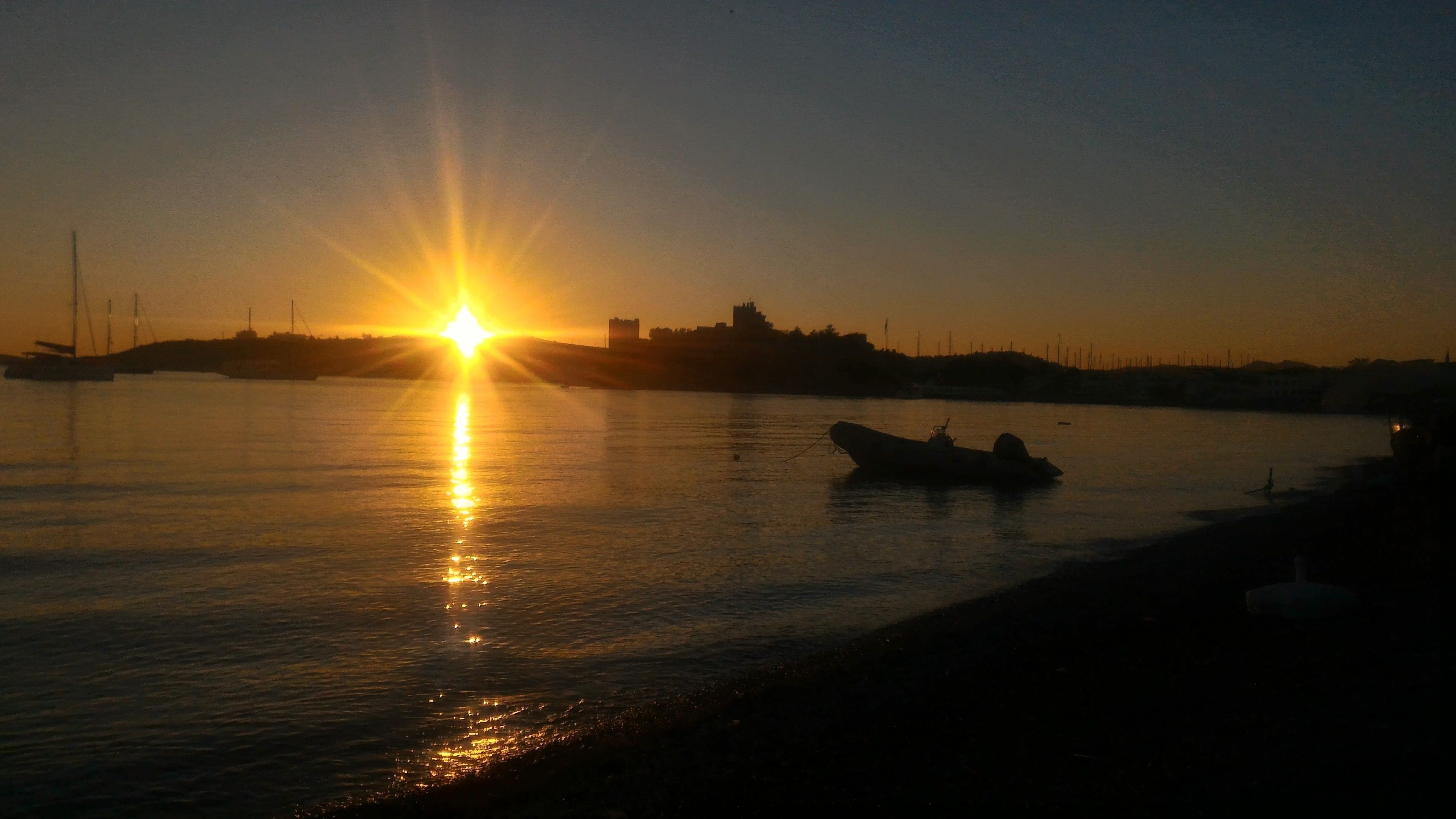 A sunset over a calm body of water with boats and distant buildings.