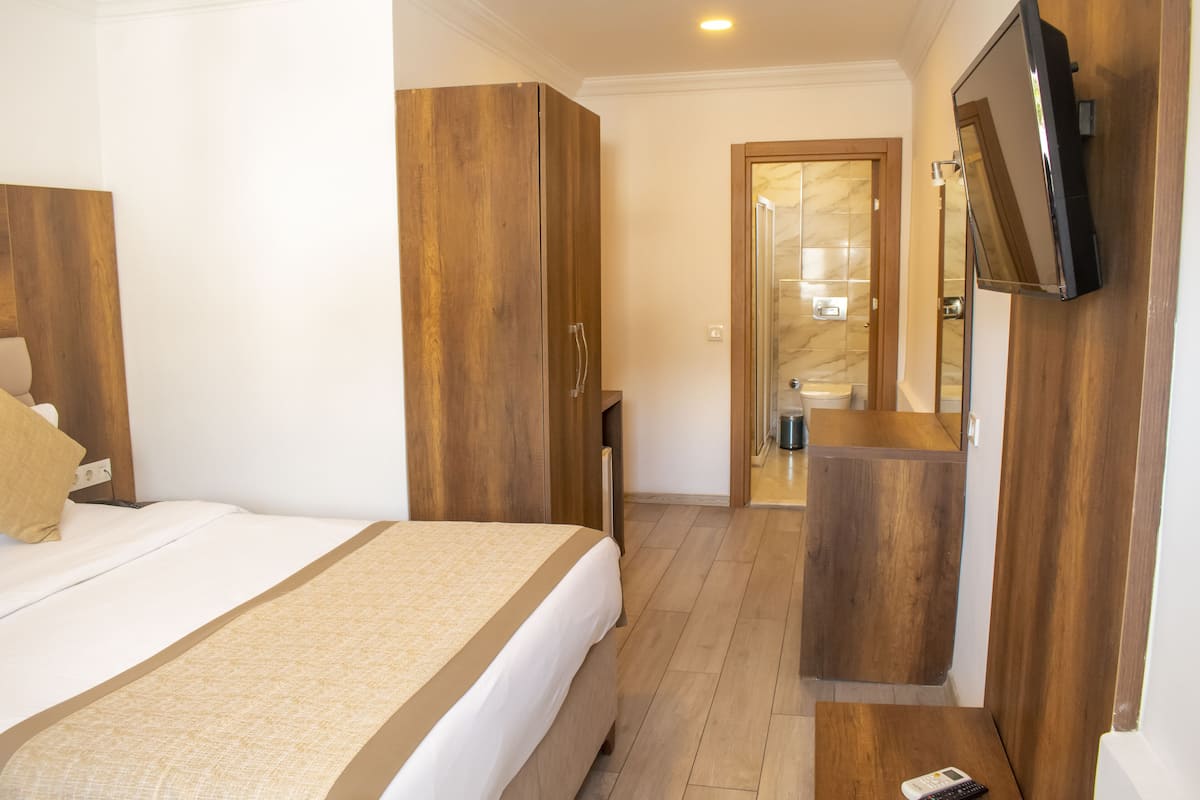 Ein Hotelzimmer mit Bett, Holzschrank, Badezimmer mit Spiegel und Waschbecken sowie einem an der Wand befestigten Fernseher.
