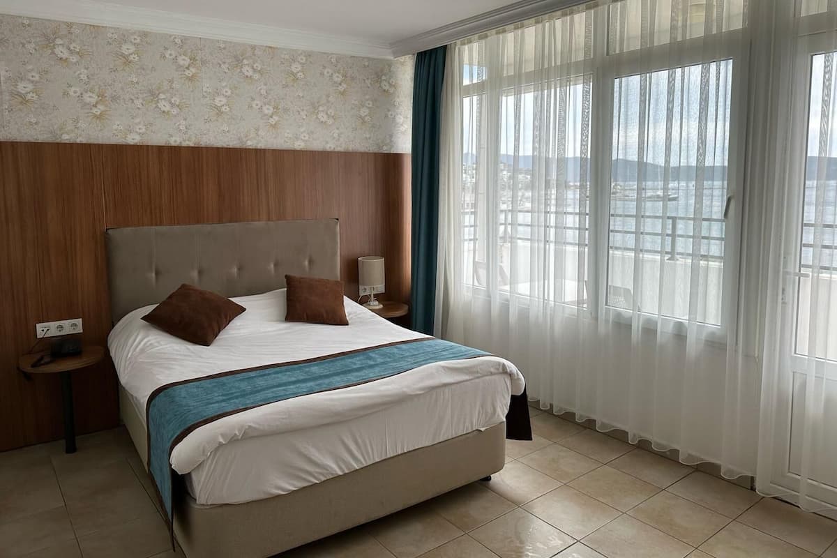 Ein Hotelzimmer mit Bett, Nachttisch, Lampe und Meerblick durch große Fenster.