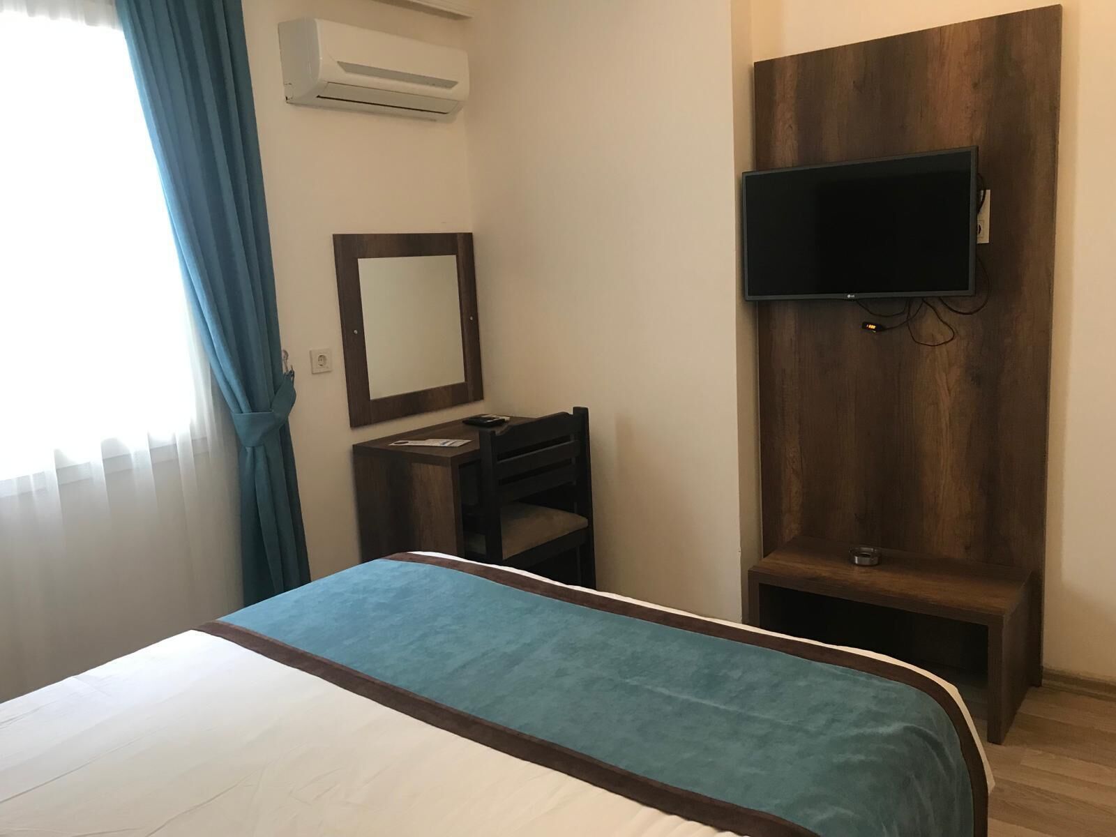 A hotel room with a bed, a desk, a chair, a TV mounted on a wooden panel, and an air conditioning unit.