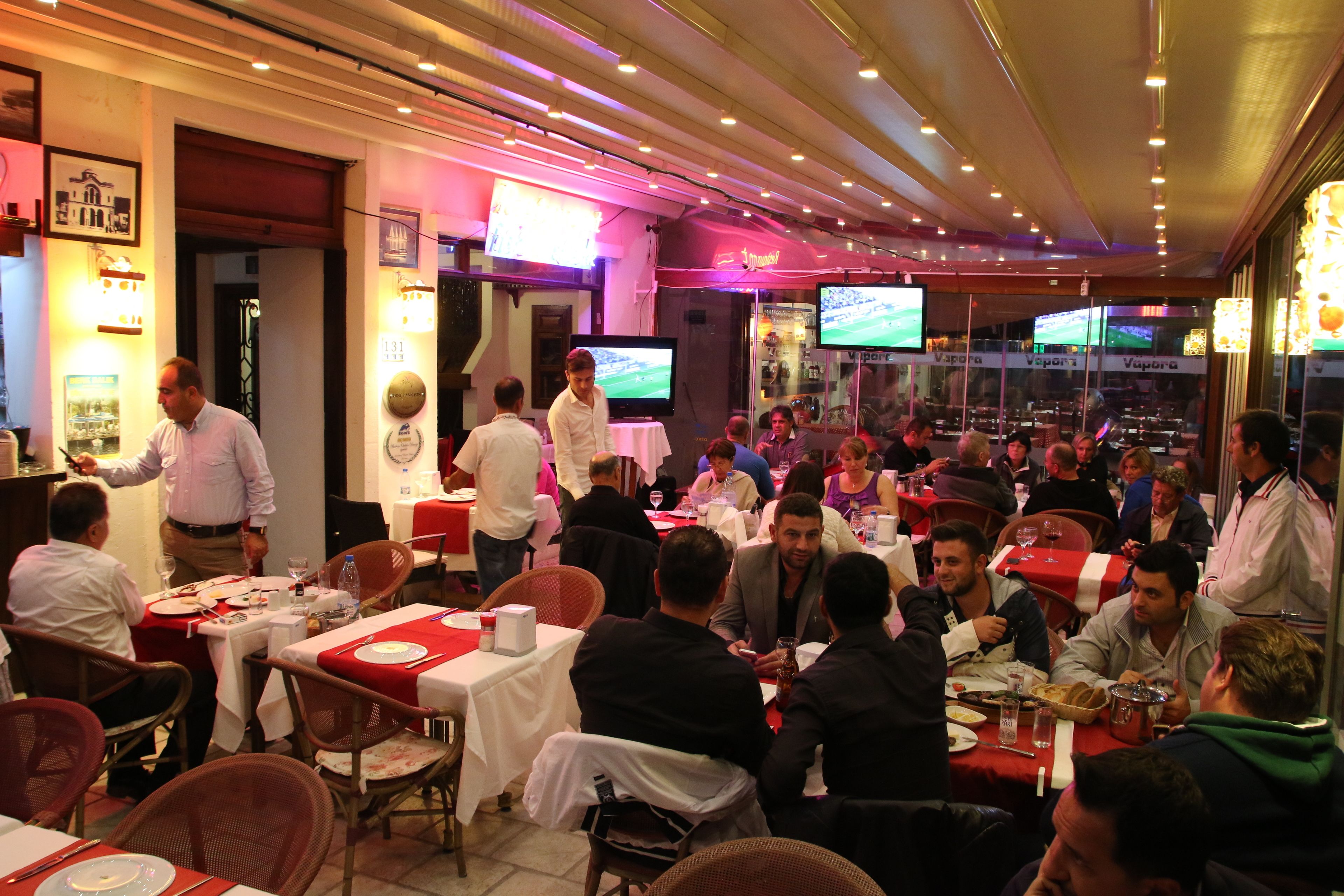 A restaurant with multiple tables and chairs, a waiter serving guests, and televisions on the wall.