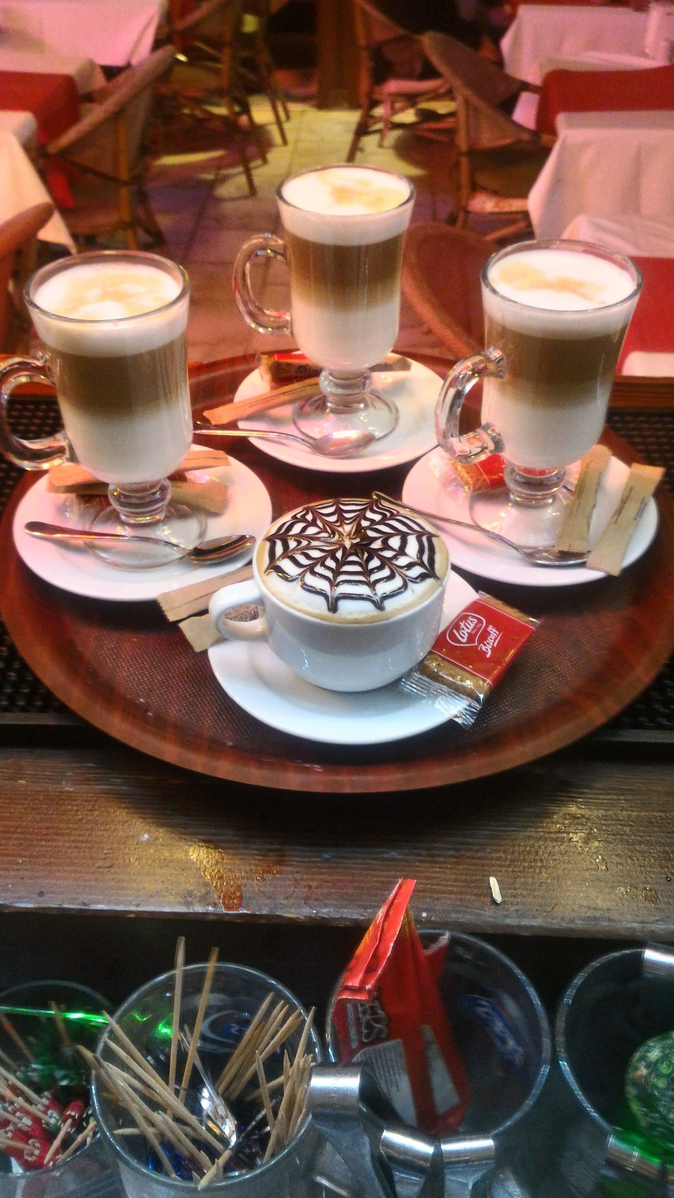 En bakke med tre kopper kaffe, hvoraf den ene har et dekorativt mønster i skummet, samt en pakke småkager.