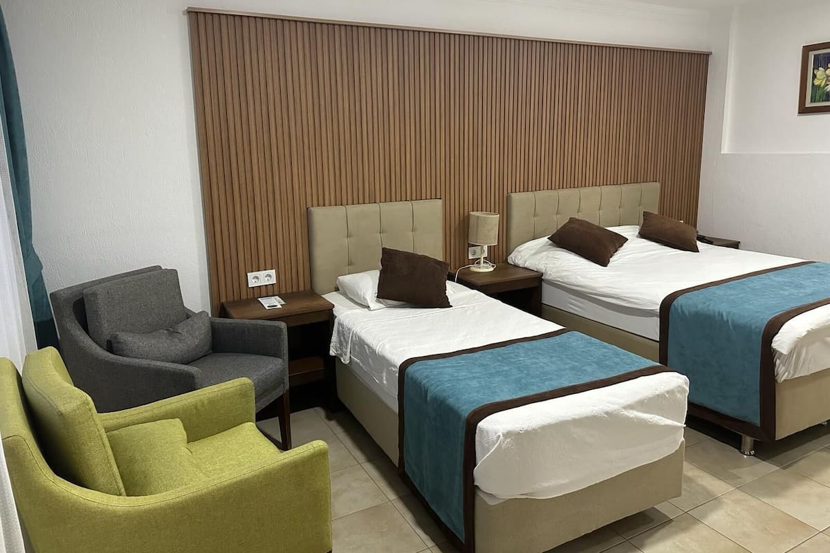 Ein Hotelzimmer mit zwei Betten, einem grünen Sessel, einem grauen Sessel und einer Wandpaneele aus Holz.