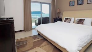 Una habitación de hotel con una cama grande, televisión, escritorio con lámpara, silla y vista al mar.
