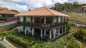 Ein zweistöckiges Gebäude mit einem Balkon, umgeben von üppiger Vegetation und blauem Himmel.