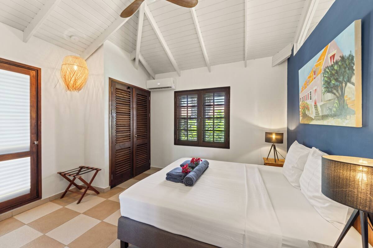 A hotel room with a bed, a window with shutters, a wall-mounted lamp, and a painting.