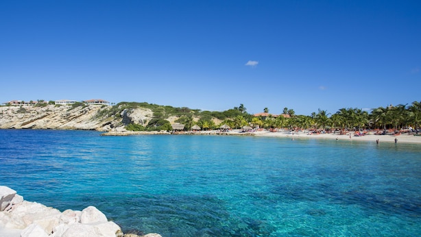 A coastal beach with clear blue water, rocky shoreline, and a line of buildings in the background. - Blue Bay Villas (Sint Michiel)