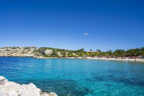 Une plage côtière bordée d’une eau d’un bleu limpide, d’une côte rocheuse et d’une rangée de bâtiments en arrière-plan.