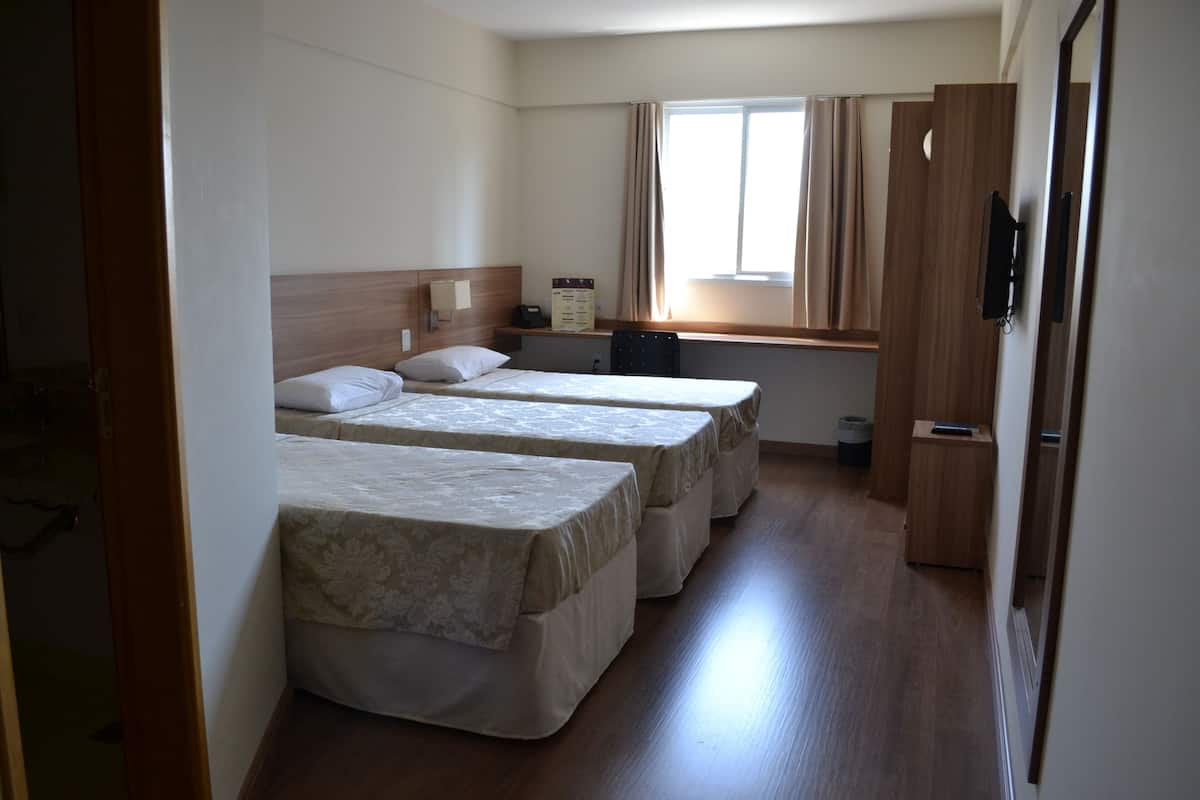 A hotel room with three beds, a wooden desk, a chair, a window with curtains, and a television on the wall.