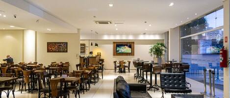 A spacious indoor area with multiple seating arrangements, including tables and chairs, a black leather sofa, and a television screen mounted on the wall.