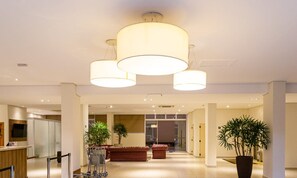 Um hall de hotel moderno com três grandes lustres cilíndricos, um balcão de recepção, uma área de estar com um sofá vermelho e plantas em vasos.