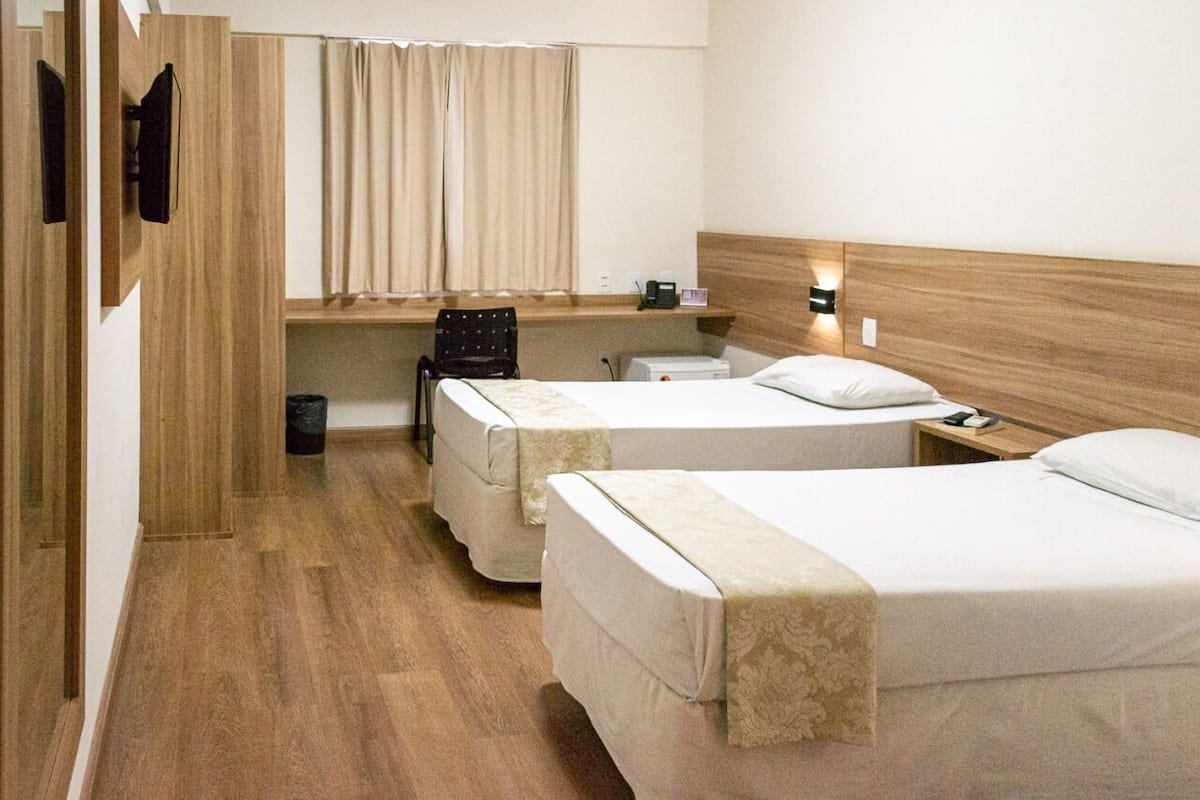 A hotel room with two beds, a desk, a chair, a small table, a window with curtains, and a wall-mounted TV.