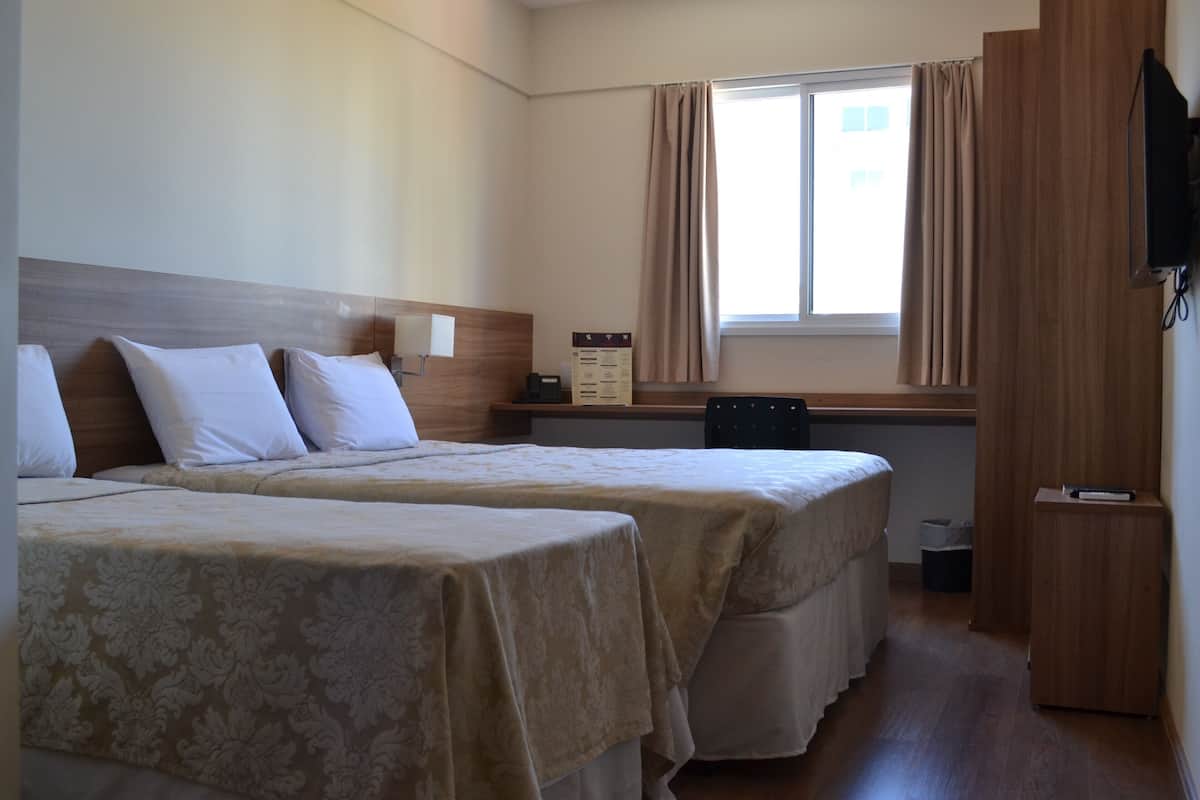 A hotel room with two beds, a wooden headboard, a window with curtains, a small table, and a television mounted on the wall.