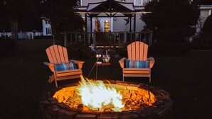 Un braciere con due poltrone Adirondack, un tavolo con bevande e una casa sullo sfondo.