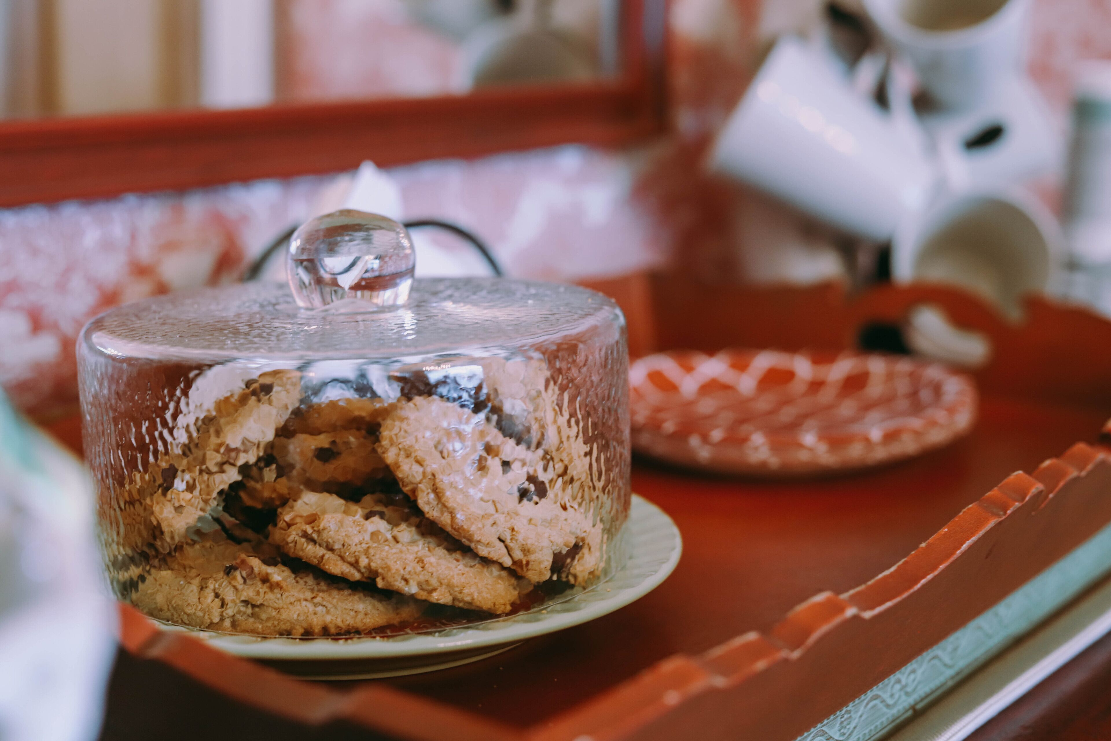 A glass-covered container with cookies on a plate, a red tray, and a white mug in the background.