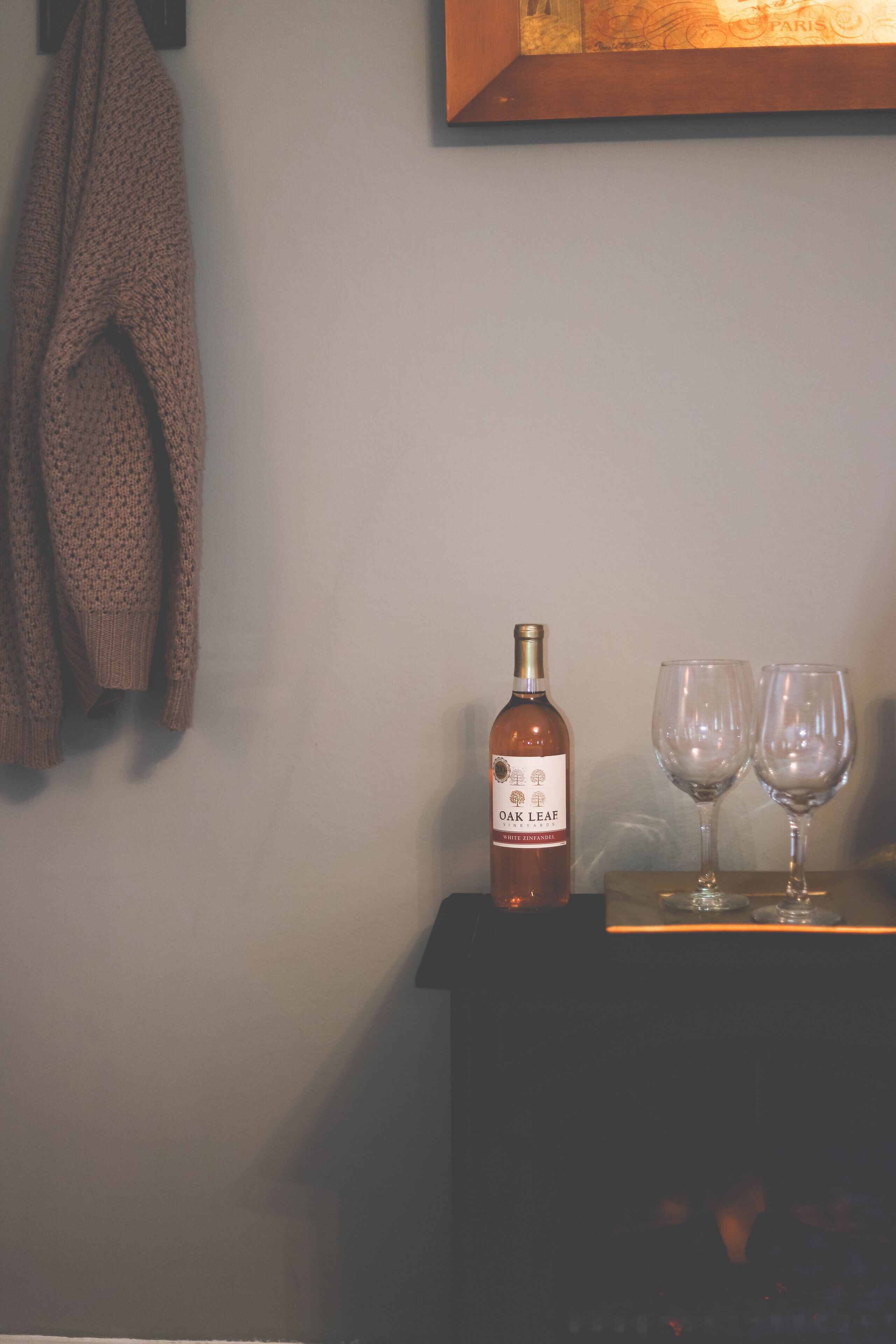 A bottle of Oak Leaf wine and two glasses on a table against a wall with a framed picture.