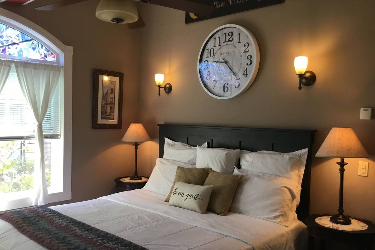 A neatly made bed with a patterned throw, two bedside lamps, a wall clock, and a framed picture.