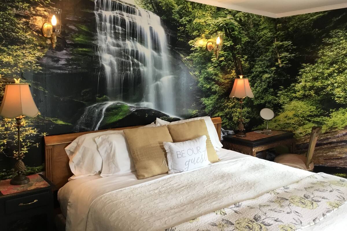 A bedroom with a large mural of a waterfall, two bedside lamps, a bed with multiple pillows, and a nightstand with a lamp.