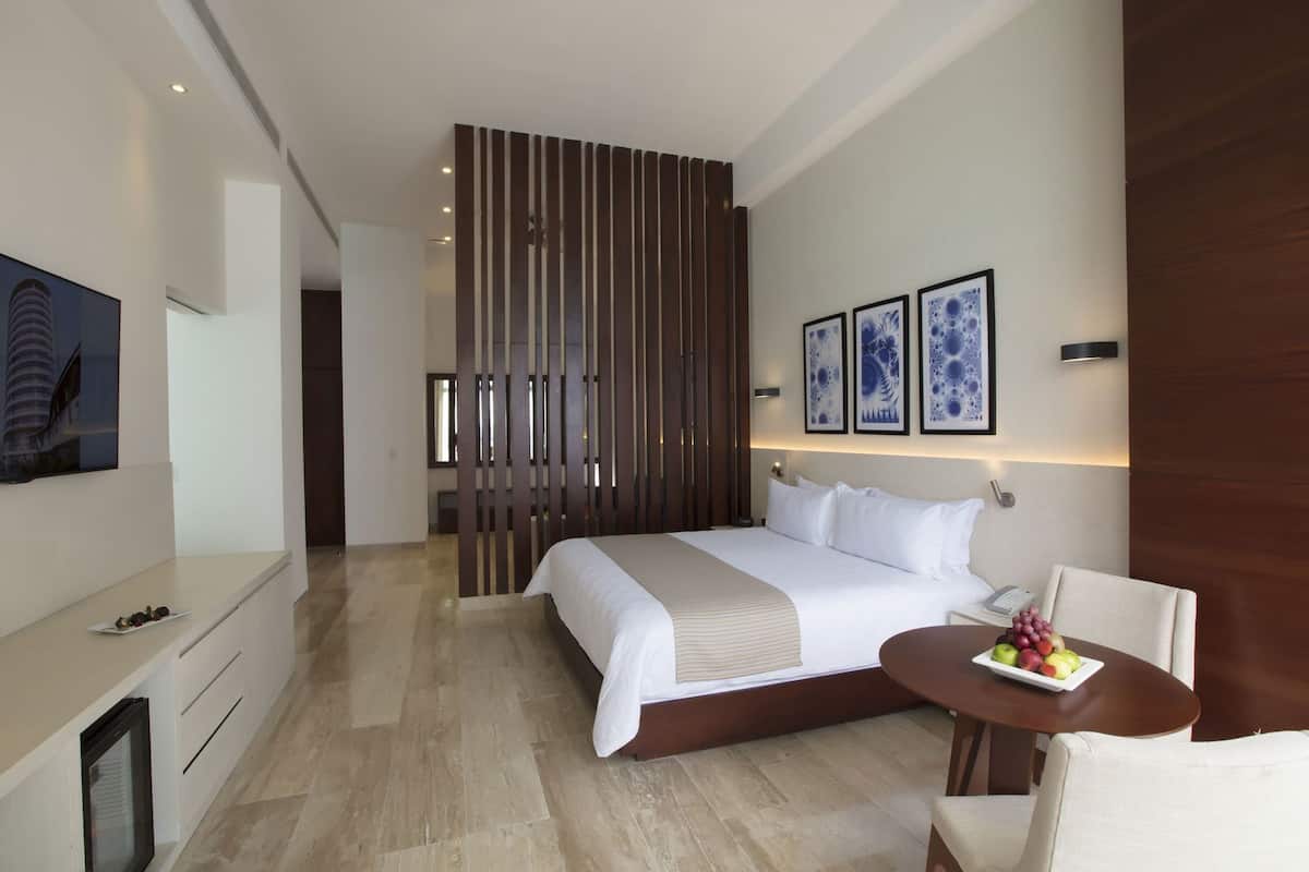 A modern hotel room with a large bed, a desk, a chair, and a small table with fruit.