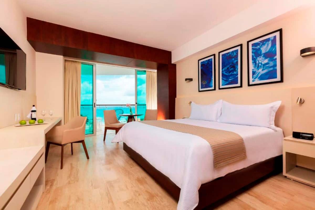 A hotel room with a large bed, a desk with a chair, a small table with a chair, and a balcony with a view.