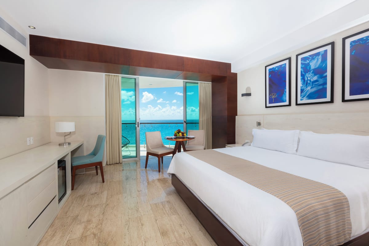 A hotel room with a large bed, a desk, a chair, and a table with fruit. There are three framed pictures on the wall and a view of the ocean through the windows.
