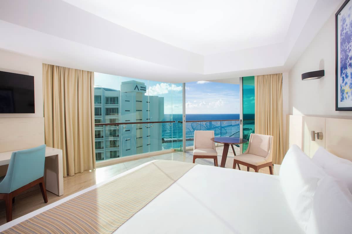 A hotel room with a large bed, a desk, two chairs, and a balcony with a view of the ocean and buildings.