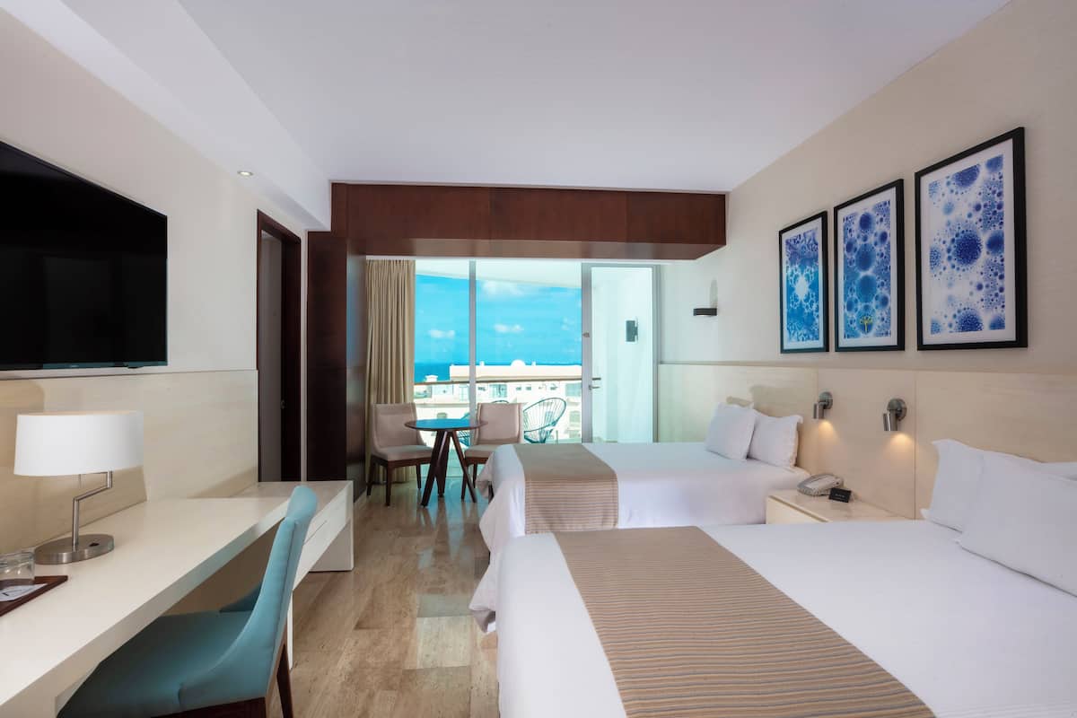 A hotel room with two beds, a desk with a lamp, a chair, a TV, and a view of the ocean.
