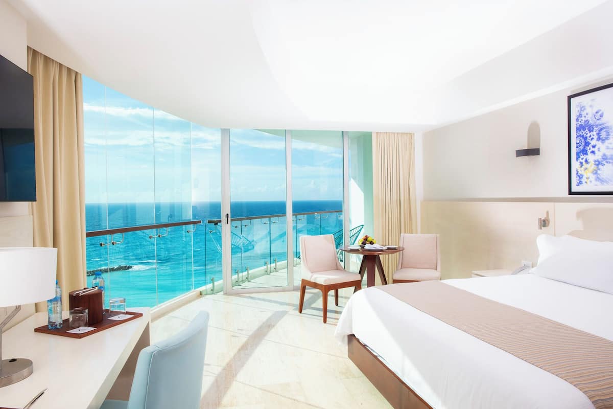 A hotel room with a large bed, a desk with a chair, a small table with chairs, and a balcony with a view of the ocean.
