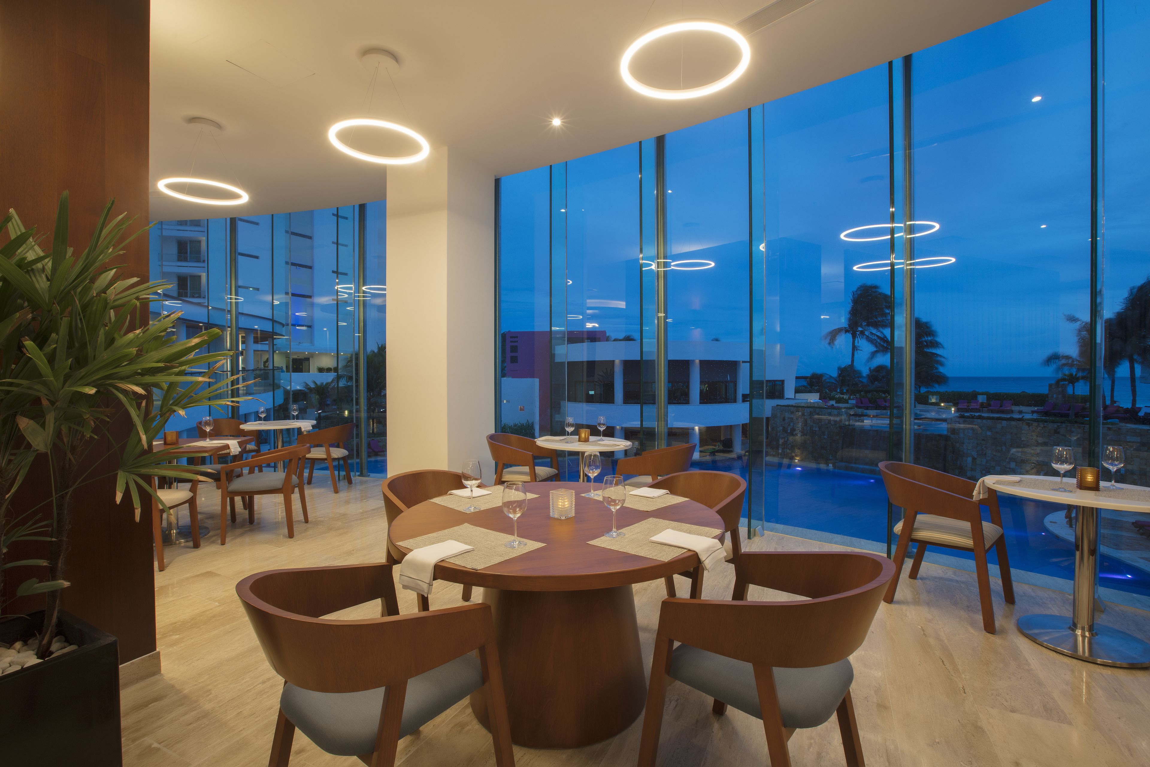 A modern dining area with round tables and chairs, large windows with a view of the pool and buildings, and circular ceiling lights.
