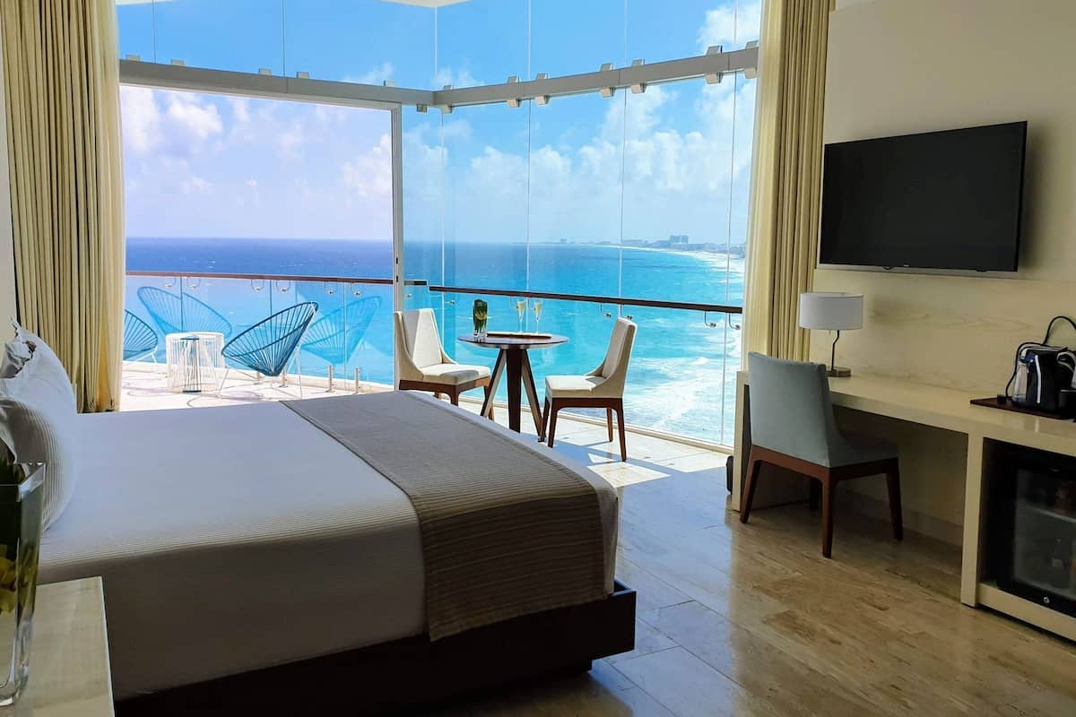 A hotel room with a large bed, a desk, a TV, and a balcony with a view of the ocean.